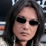 Tomonobu Itagaki, the creator of Dead or Alive and Ninja Gaiden, with his signature long black hair and silver-rimmed sunglasses, looking directly ahead.