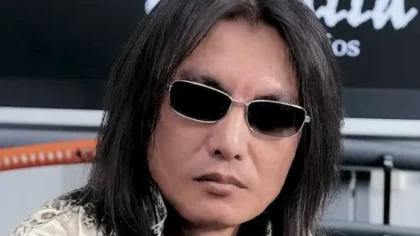 Tomonobu Itagaki, the creator of Dead or Alive and Ninja Gaiden, with his signature long black hair and silver-rimmed sunglasses, looking directly ahead.