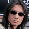 Tomonobu Itagaki, the creator of Dead or Alive and Ninja Gaiden, with his signature long black hair and silver-rimmed sunglasses, looking directly ahead.