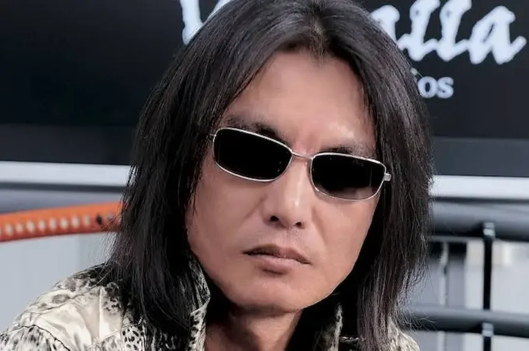 Tomonobu Itagaki, the creator of Dead or Alive and Ninja Gaiden, with his signature long black hair and silver-rimmed sunglasses, looking directly ahead.